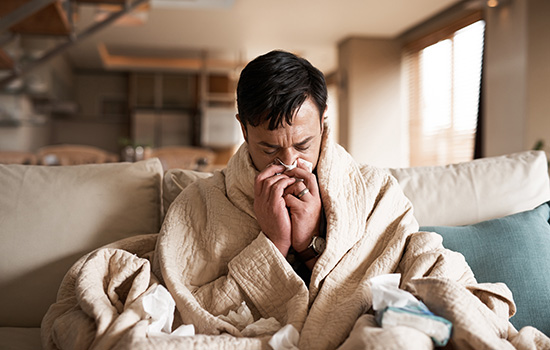 A young white man on his couch, wrapped in blankets, blowing his nose. Tissues are sprinkled around him.