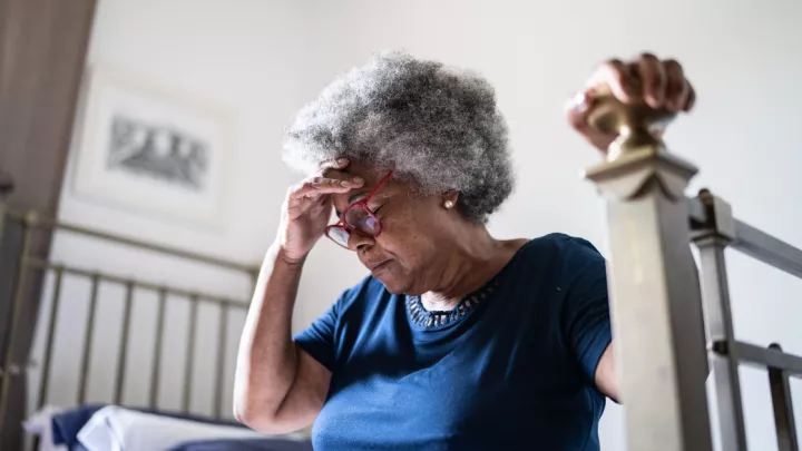 A picture of an older woman sitting on her bed holding her head